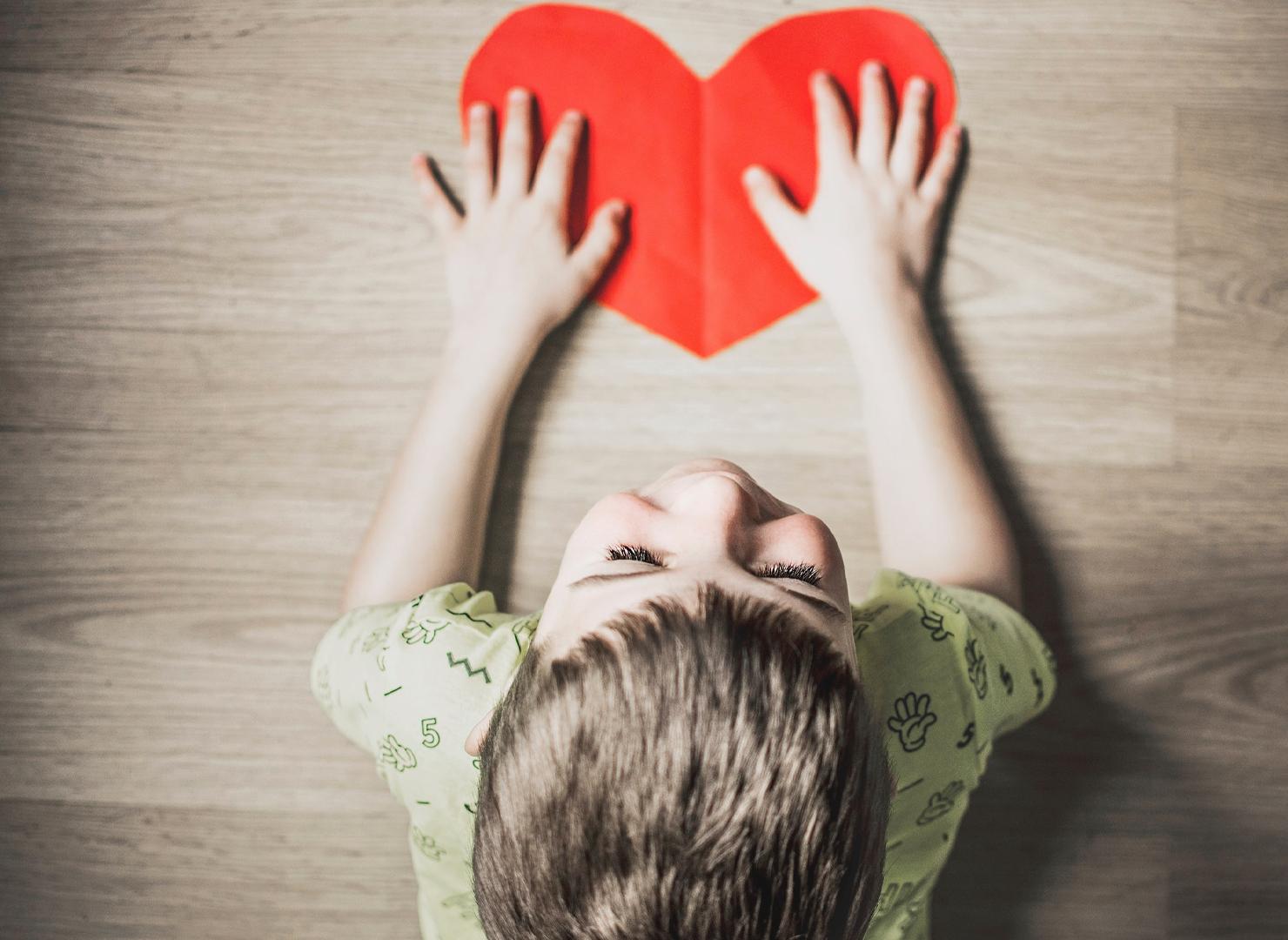 A young boy placing his hands over a red paper heart on a table in front of him.